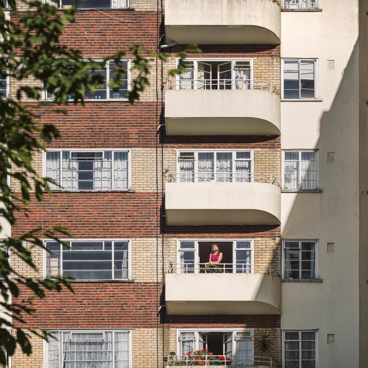 Photograph of an Art-Deco style 1930’s residential building, with alternating red and yellow brick floors.  Captured with a long lens from a distance.  On second balcony from the bottom there is a woman in a red blouse standing with her hands on the railings.  A hard shadow cast by the sun covers one third of the building.  The canopy of a tree partly obscures the left side of the frame.