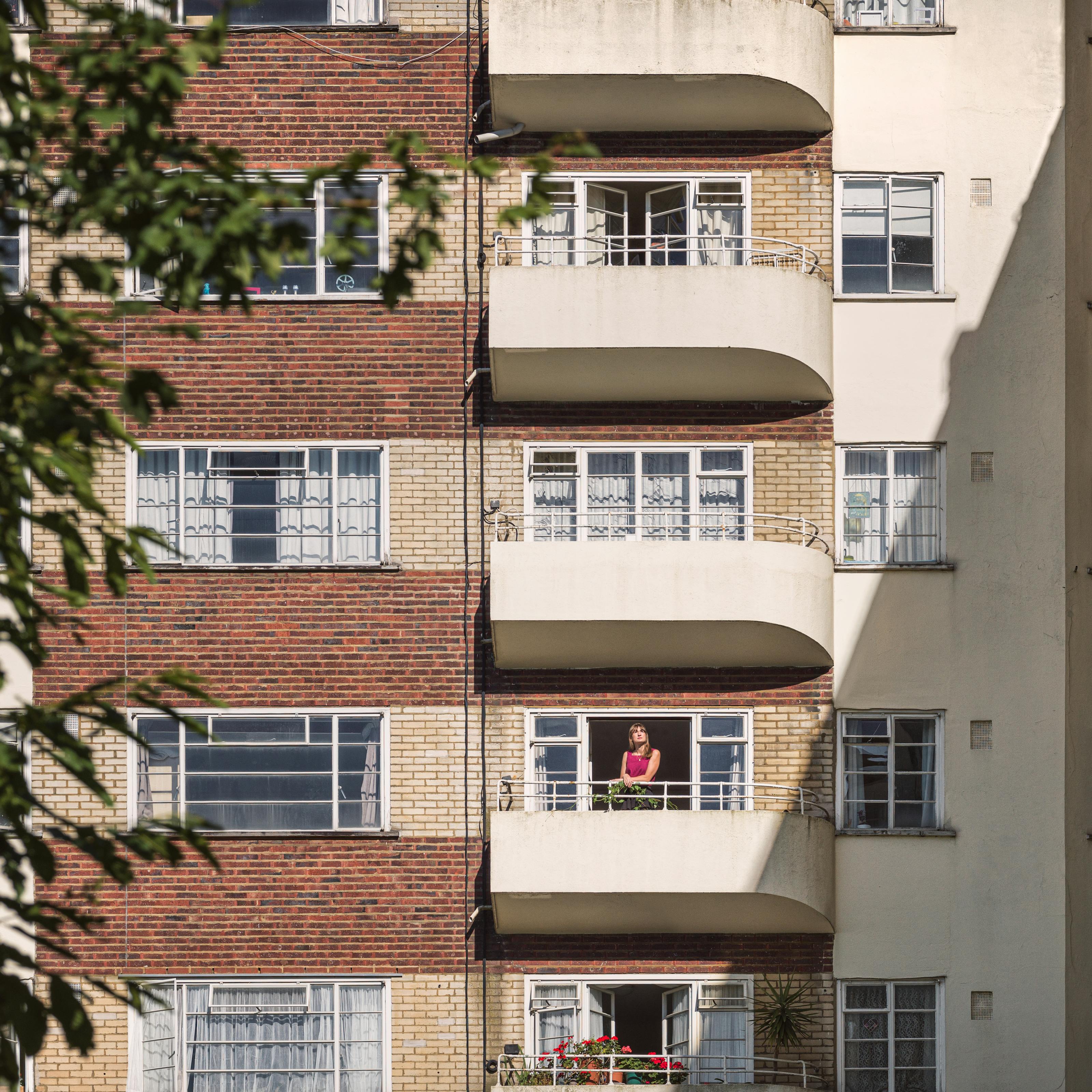 Photograph of an Art-Deco style 1930’s residential building, with alternating red and yellow brick floors. Captured with a long lens from a distance. On second balcony from the bottom there is a woman in a red blouse standing with her hands on the railings. A hard shadow cast by the sun covers one third of the building. The canopy of a tree partly obscures the left side of the frame.