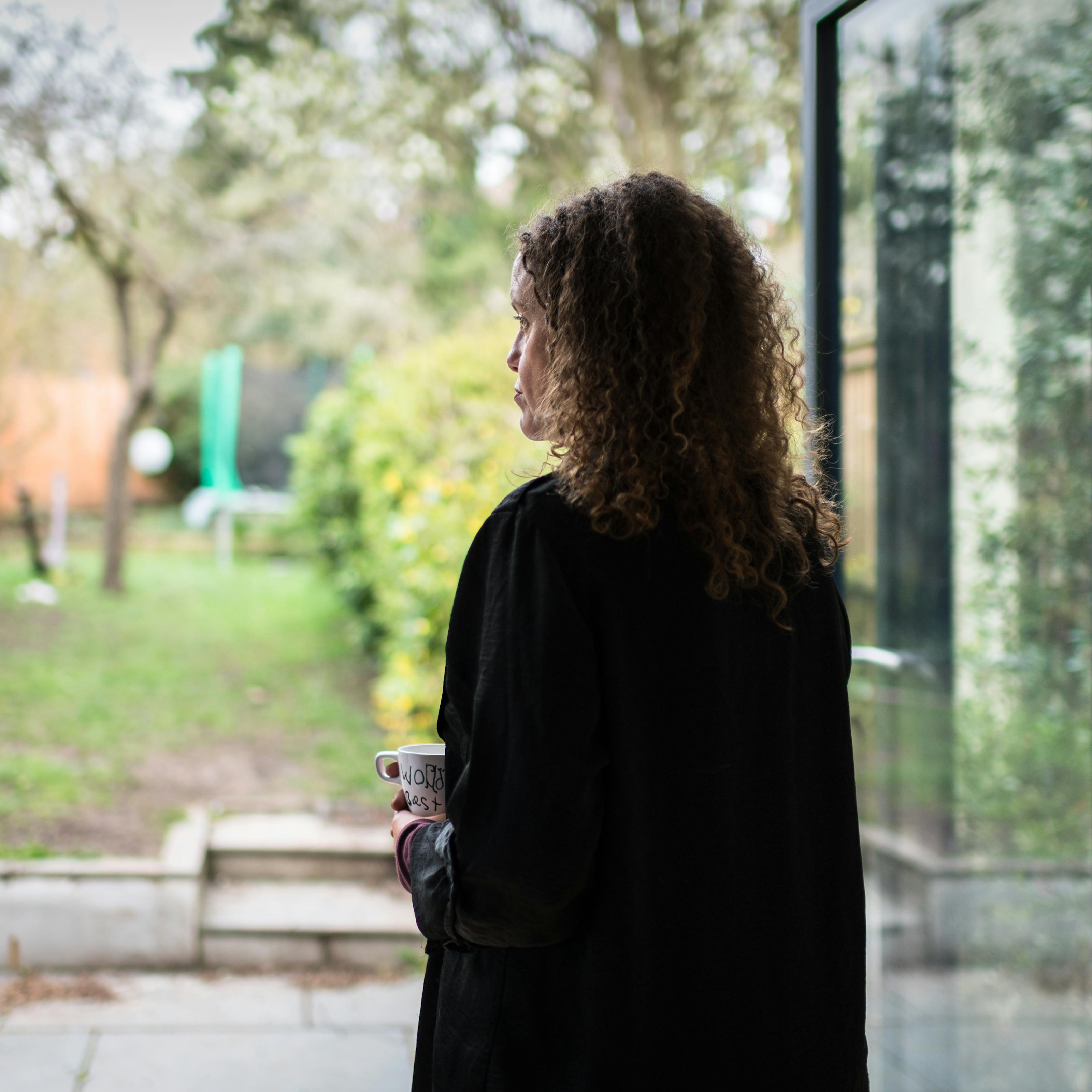 Photograph of a woman standing next to a set of bifold doors looking out into a domestic garden. The left profile of her face can been seen looking into the distance. She is holding a mug.