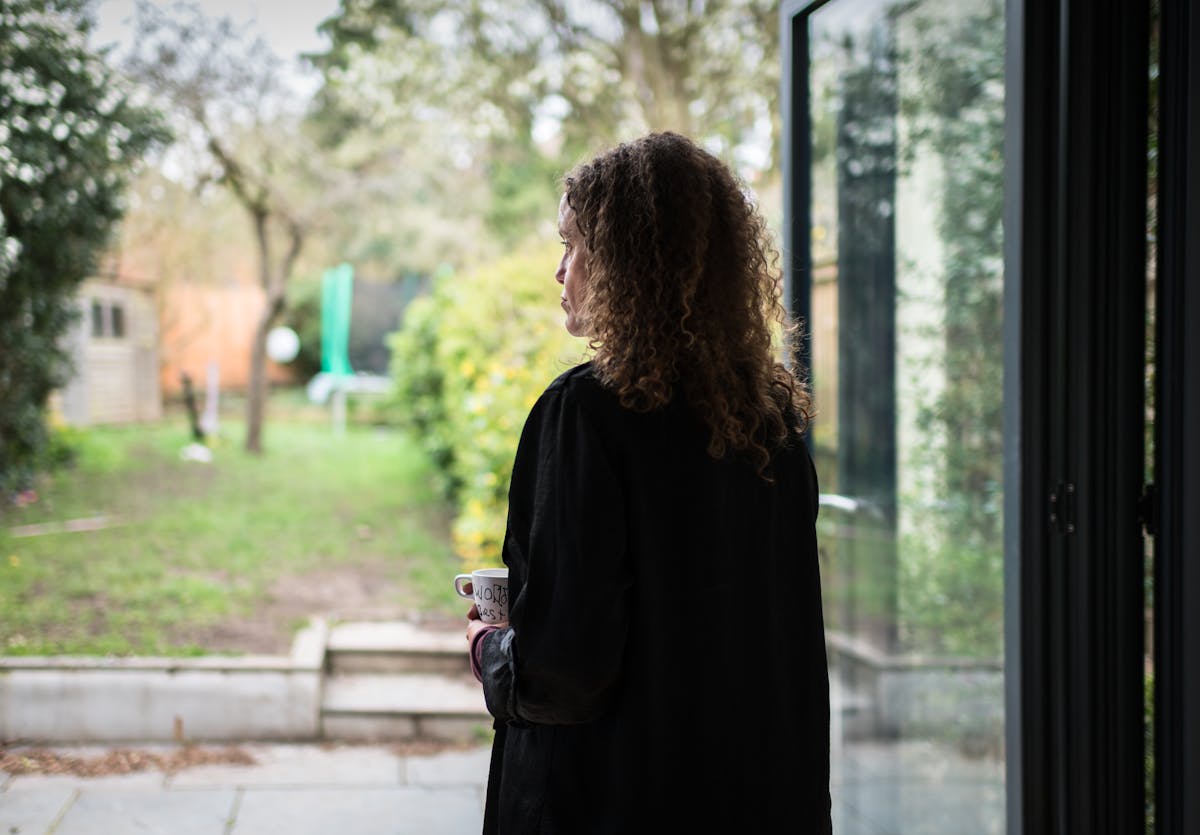 Photograph of a woman standing next to a set of bifold doors looking out into a domestic garden. The left profile of her face can been seen looking into the distance. She is holding a mug.