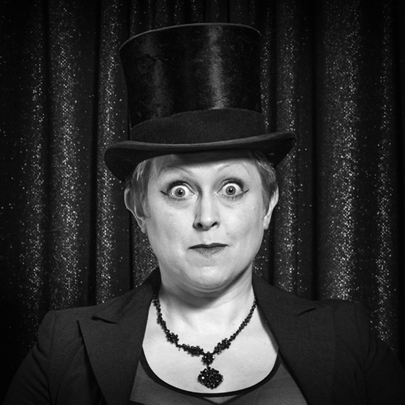 Photographic black and white, head and shoulders portrait of Naomi Paxton in a top hat against a glittery background.