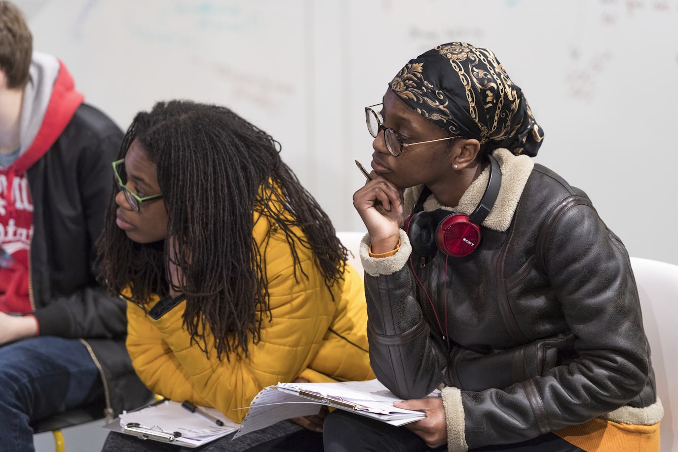 Two young people sit leaning forward attentively listening, with writing paper on their laps and pens in hand.