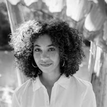 Black and white photograph showing Delphine Sims from the bust up, wearing a white button upped, shirt. Her curly hair falls around her full face as she looks up at the camera slightly smiling. Just above her head is a large tropical leaf and behind her is a blurred background of water and tree trunks. 