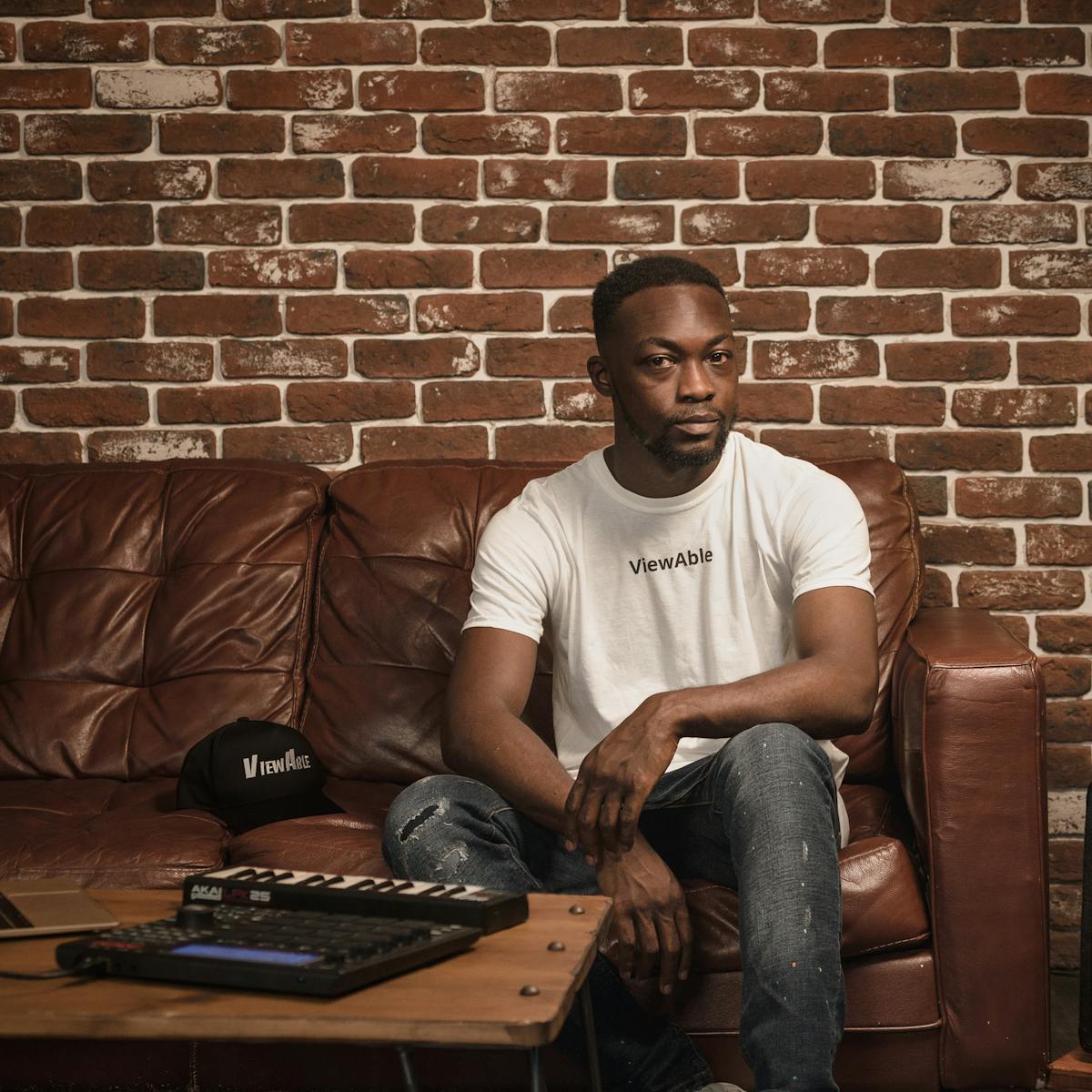 Photographic full length portrait of a man in a white t-shirt sitting on a leather sofa. In front of him to the left of the frame is a wooden table with a laptop, keyboard and beat-box. To the right of the frame is a bass speaker and a floor standing lamp.  Beside him on the sofa is a hat. A single line of text is legible on both the T-shirt and hat, reading 'Viewable'.