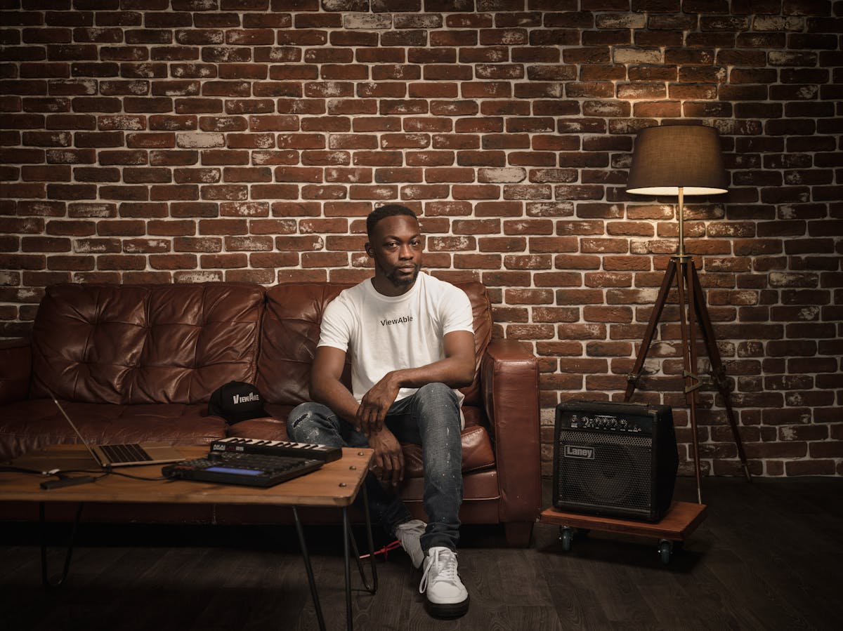 Photographic full length portrait of a man in a white t-shirt sitting on a leather sofa. In front of him to the left of the frame is a wooden table with a laptop, keyboard and beat-box. To the right of the frame is a bass speaker and a floor standing lamp.  Beside him on the sofa is a hat. A single line of text is legible on both the T-shirt and hat, reading 'Viewable'.