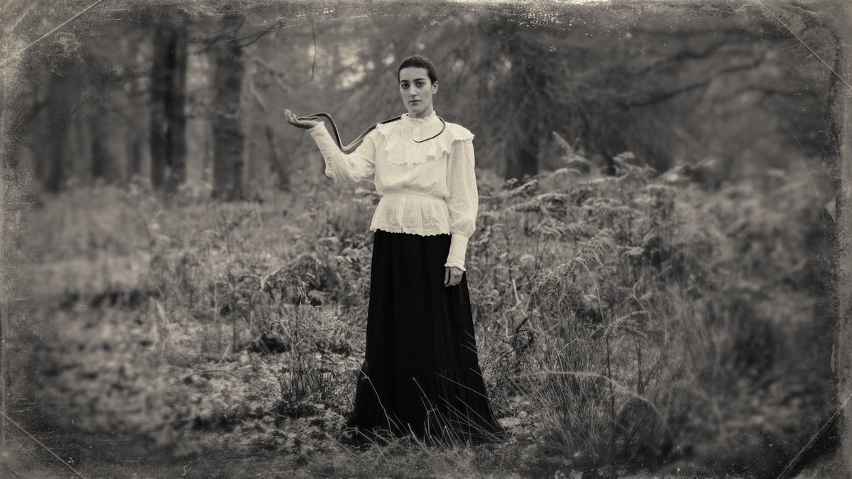 Sepia toned photograph with a digital filter applied to give the impression the photograph was created using a glass-plate process from the mid 19th century. The effect of this filter includes scratches and fingerprints. The photograph shows a woodland scene made up of tall grasses, bracken and distant trees. In the centre of the image stands a tall woman dressed in a black floor length skirt and a white blouse embellished with frills and large cuffs. She is looking straight to camera, her right arm is raised to her side, palm facing upwards. Wrapped around her neck and running over her shoulder and down to her open hand is a long dark toned snake. The centre of the frame is in sharp focus, but the edges descend quickly into a blur.