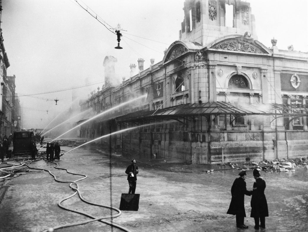 Black and white photograph showing water jetting out of hose pipes onto a market building. Police and fire crew in the foreground.