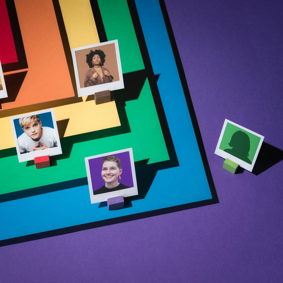 Photograph of four portraits featuring LGBTQ+ comedians, framed within a white paper frames.  To the right of the image, an anonymous figure is also framed within a white frame. Layered beneath them in tiers are the LGBTQ+ colours of red, orange, yellow, green blue and purple; which forms the floor of the set.  The colours are laid out in such a way as they form a chevron.