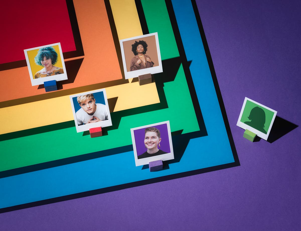 Photograph of four portraits featuring LGBTQ+ comedians, framed within a white paper frames.  To the right of the image, an anonymous figure is also framed within a white frame. Layered beneath them in tiers are the LGBTQ+ colours of red, orange, yellow, green blue and purple; which forms the floor of the set.  The colours are laid out in such a way as they form a chevron.