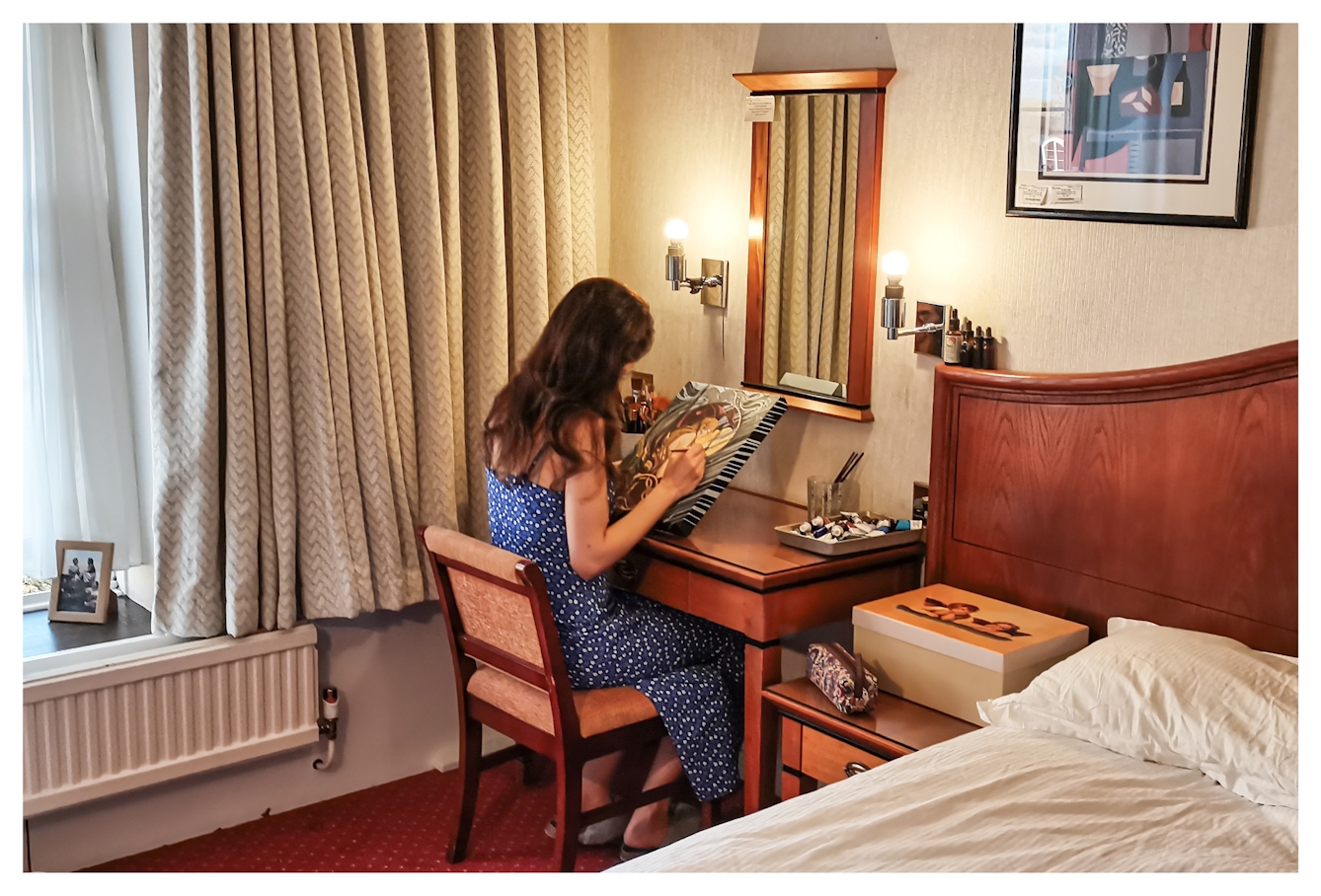 Photograph of the inside of a hotel room, with a corner of the bed, window, curtains and a wall mirror. Sat at the small desk is a woman in the process of painting onto a small stretched canvas.