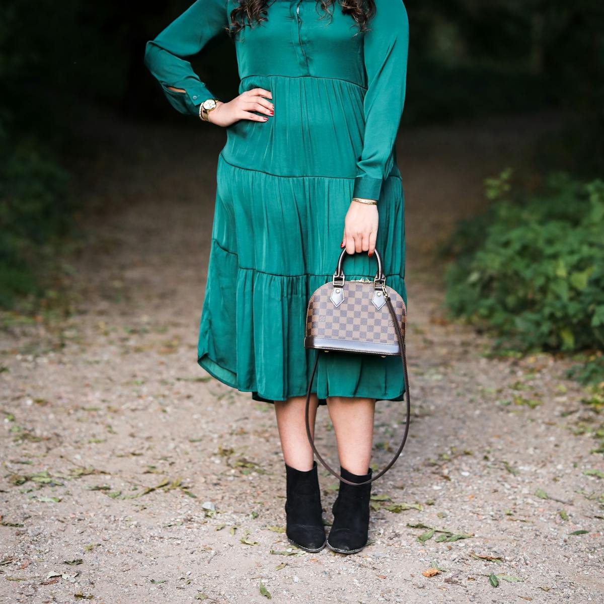 Photograph of a woman from her shoulders to her feet, wearing an emerald green dress with a shiny lustre. She is standing with her right hand on her hip and in her left hand she is holding a small hand bag in front of her left leg. In the background can be seen a park or woodland scene with a footpath disappearing into the distance. On either side of the path is green vegetation.
