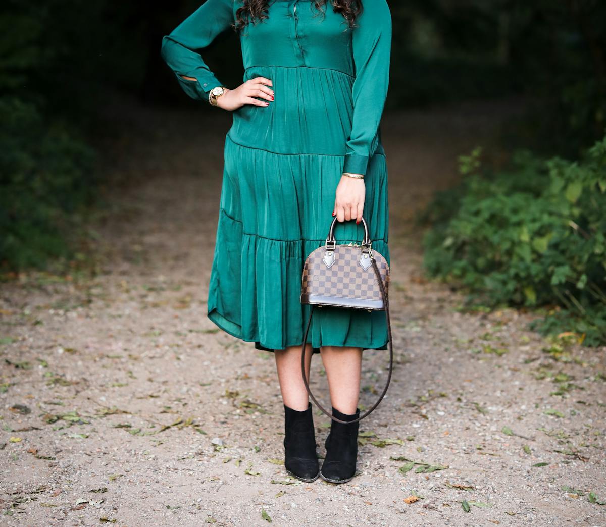 Photograph of a woman from her shoulders to her feet, wearing an emerald green dress with a shiny lustre. She is standing with her right hand on her hip and in her left hand she is holding a small hand bag in front of her left leg. In the background can be seen a park or woodland scene with a footpath disappearing into the distance. On either side of the path is green vegetation.