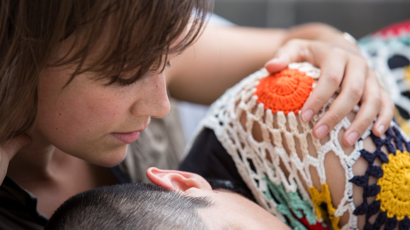 A photographic image of two people. One, with face in profile, tenderly holds the shoulder of a second, lying on their side and wearing a colourful, loosely-crocheted top.  