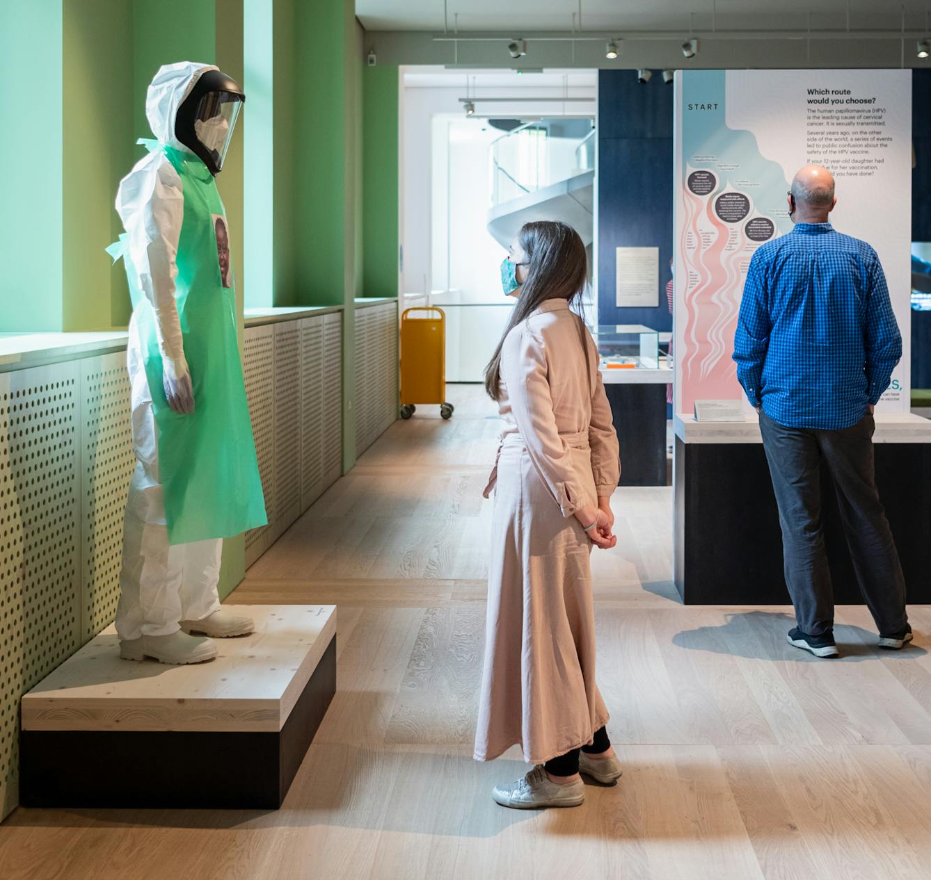 A photograph showing a visitor looking at an exhibition exhibit of a mannequin wearing a white hazmat suit, visor and green apron standing on a low wooden platform. On the green apron there is a portrait of the carer that would have been wearing the suit whilst treating Ebola patients. To the right is the back of another visitor who is looking at a different exhibit.