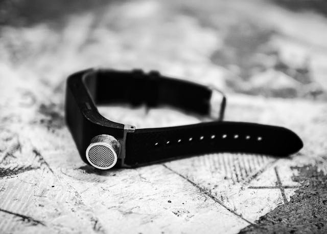 Black and white photograph of a watch like object resting on its side on a textured background. The device has a black strap and buckle. In the centre of the strap is a cylindrical metal attachment with a wire mesh front. The image has a very shallow depth of field, throwing the foreground and background out of focus.