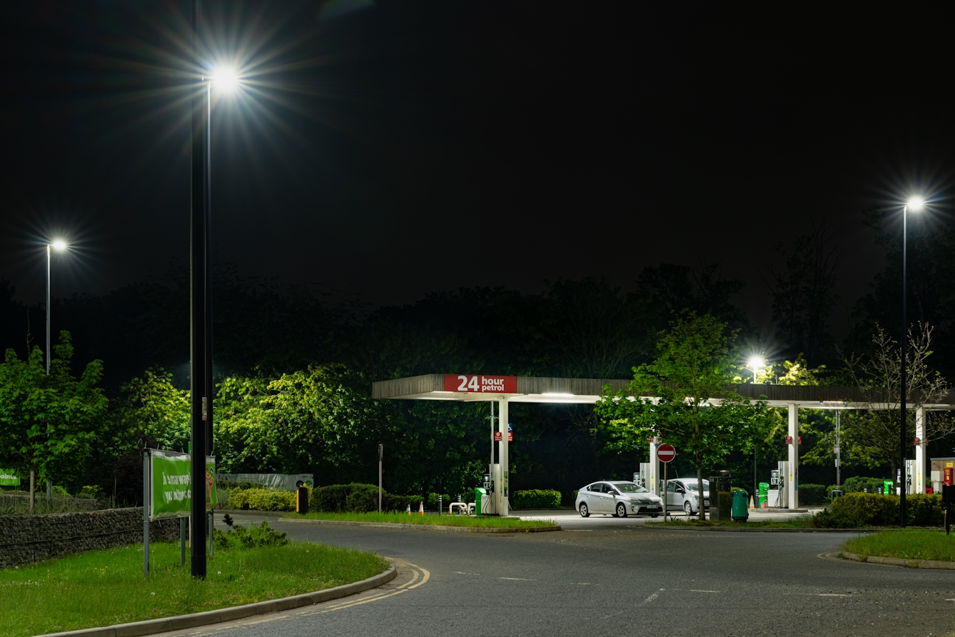 A photograph of a petrol station at night. At there petrol pumps are two cars and above there is a sign that reads 24 hour petrol. Surrounding the petrol station are streetlights, which illuminate the grass and trees in the verges next to the road.