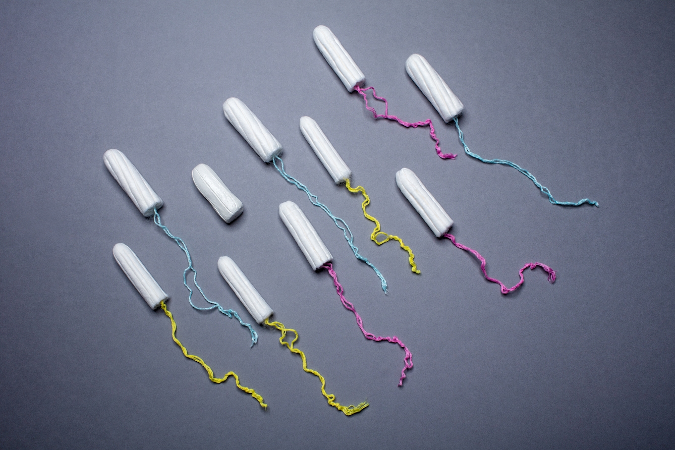 Photograph of 10 unwrapped tampons arranged in a diagonal pattern on a blue background. The yellow, pink and blue cords are trailed behind each tampon except for one.