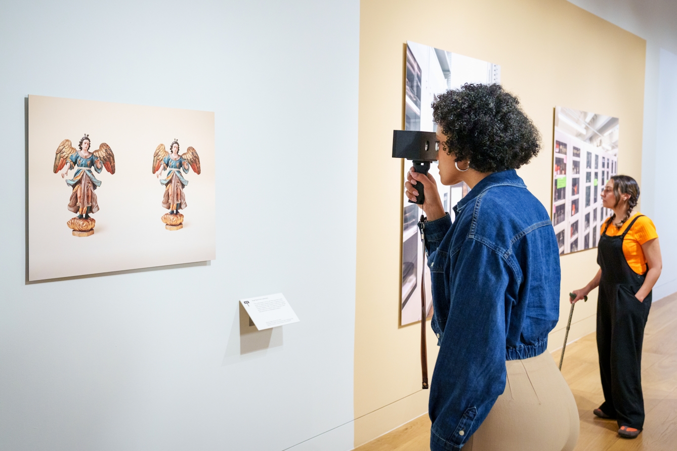 Photograph of a gallery exhibition space showing large photographic prints mounted on the walls. The colours of the room are light whites and yellows. One visitor wearing a denim shirt is holding a stereoscopic viewer to their eyes, focusing on a photographic print on the wall which depicits a ceramic angel figure, shown in duplicate. Standing in the background is another visitor in an orange t-shirt holding a walking stick who is looking any the artworks. 