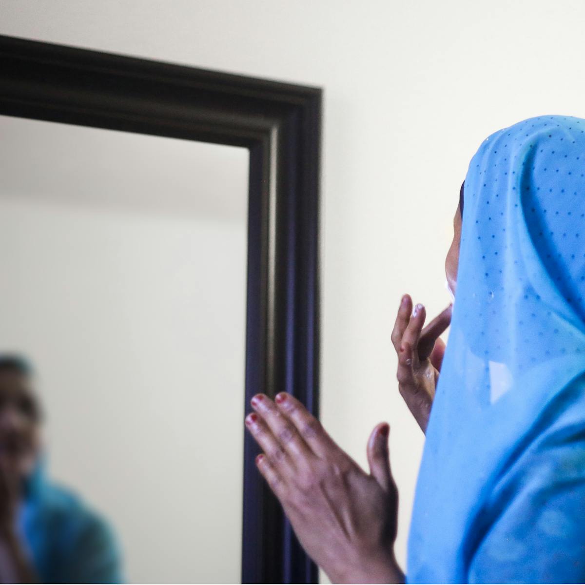 Photograph of the back of a woman's head and shoulders as she applies face cream in front of a wall mirror. In the reflection you can see her face, out of focus.