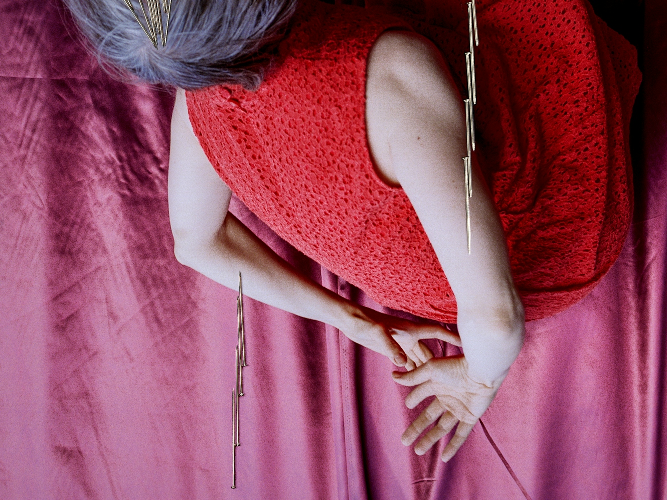 Detail from a larger artwork created with a colour photographic print of a female figure in a bright red dress, set against a purple and blue draped silk background. She is curled up in the frame with her hands loosely clasped behind her back. Her body is targeted by groups of dress pins, laid on top of the photographic print. The pins are arranged as if they are a flight of arrows directed at her body. Two group attack her arms from underneath and above.