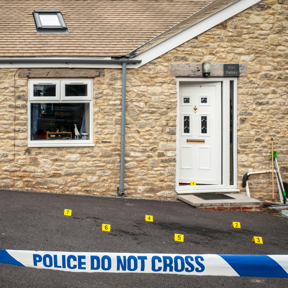 Photograph of the front of a house with the front door slightly open and forensic crime scene numbered yellow markers on the ground. In the foreground is a police cordon with 'Police Do Not Cross' written across the tape.