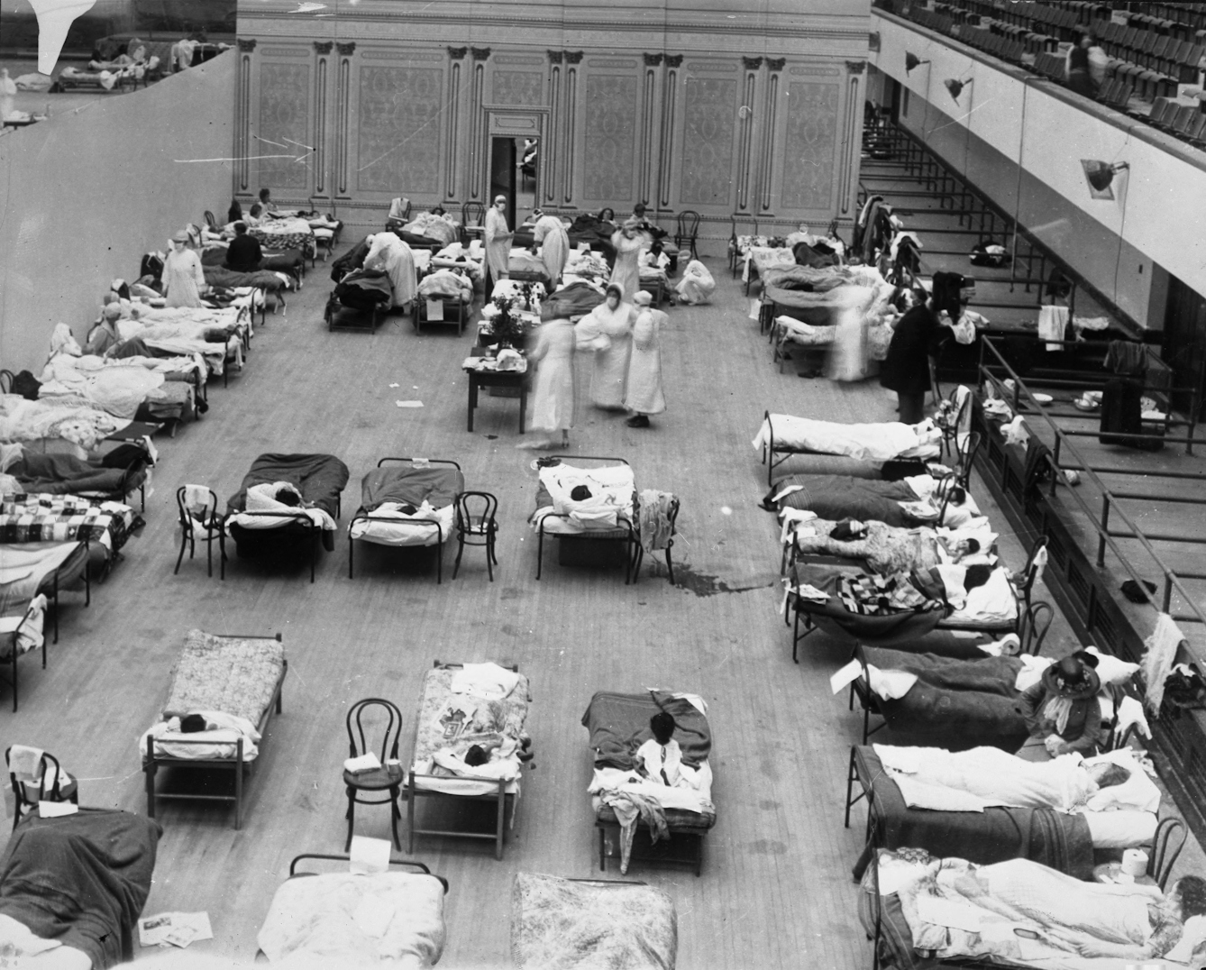 The photograph depicts volunteer nurses from the American Red Cross tending influenza sufferers in the Oakland Auditorium, Oakland, California, during the influenza pandemic of 1918.