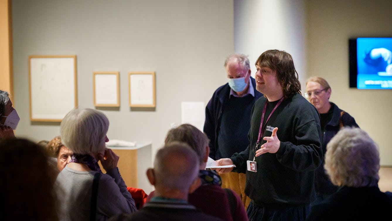 A Visitor Experience Assistant standing in a gallery talking to visitors. They are holding some notes and wearing a black jumper with 'Wellcome Collection' written on it. 