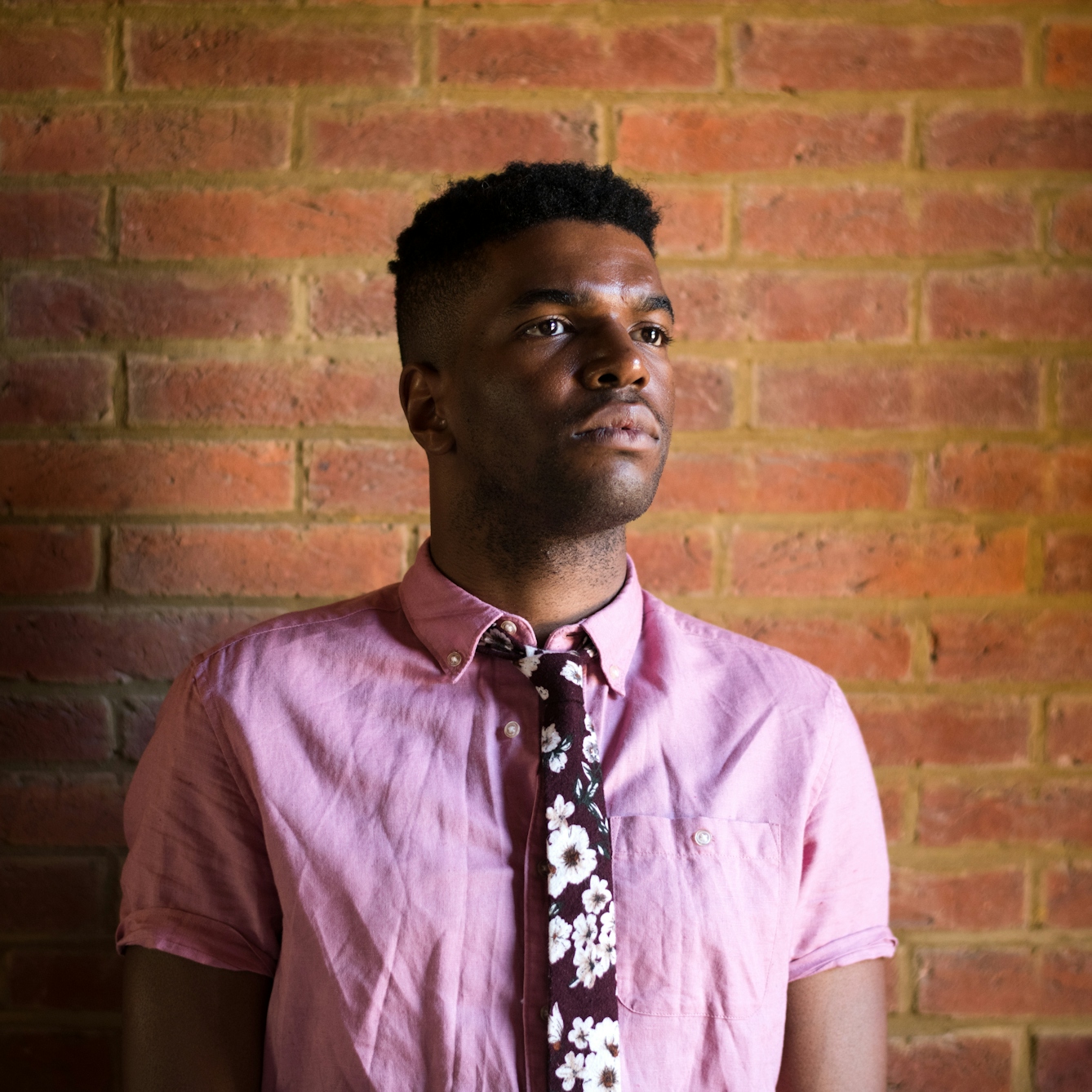 Photograph of a man wearing a pink shirt and floral tie standing in front of a red brick wall, look off camera into the distance.