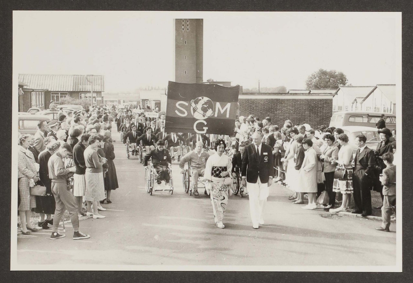 Black and white photographic print of a parade. A woman in a kimono and a man in a dark blazer and white trousers lead the parade, and behind them follow row upon row of people in wheelchairs wearing tracksuits or suits. The front row of people in wheelchairs carries a banner that says SMG with a globe above the letter G. The street is lined with people in 1950s clothing, who watch the parading athletes. One man in the front left of the photograph is wearing a tracksuit and Converse sneakers and is taking photographs.
