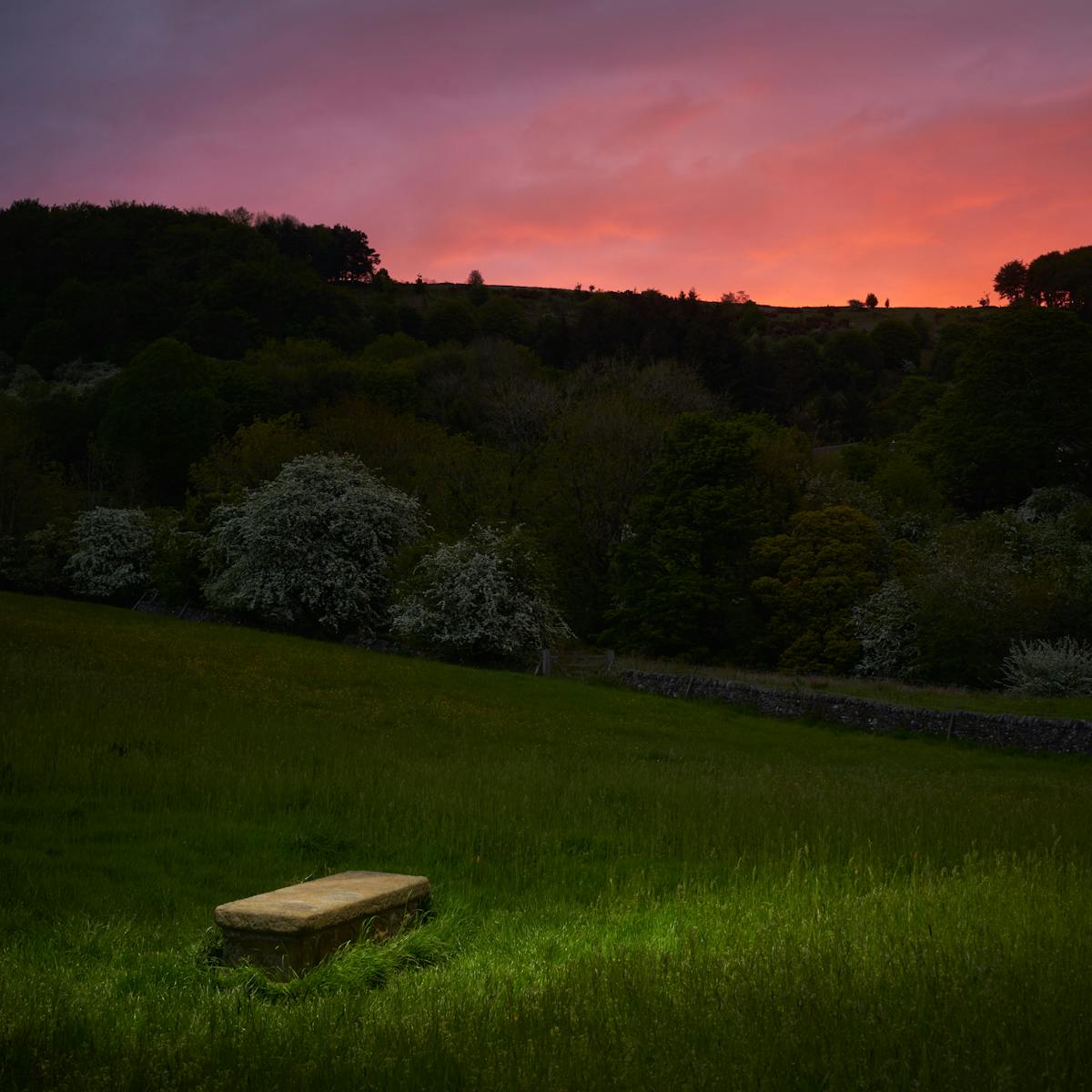 Photograph of a rural landscape at dusk. In the background is a dramatic red sky. In the foreground is a spotlit plain stone grave.