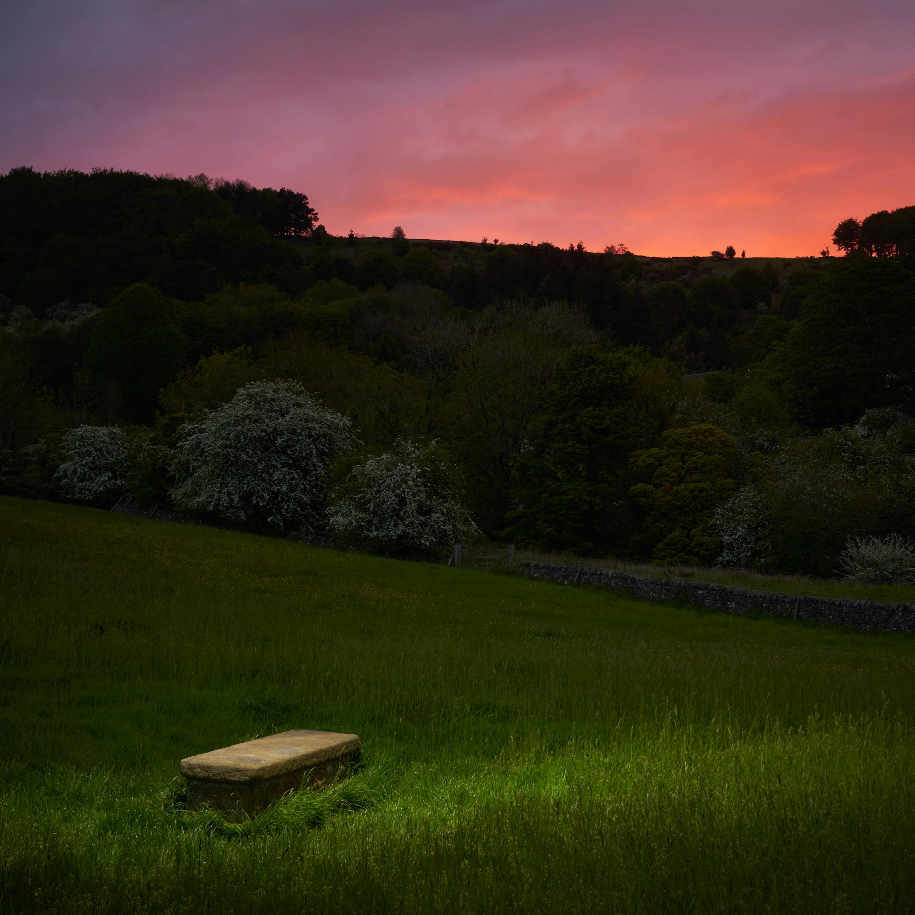 Photograph of a rural landscape at dusk. In the background is a dramatic red sky. In the foreground is a spotlit plain stone grave.
