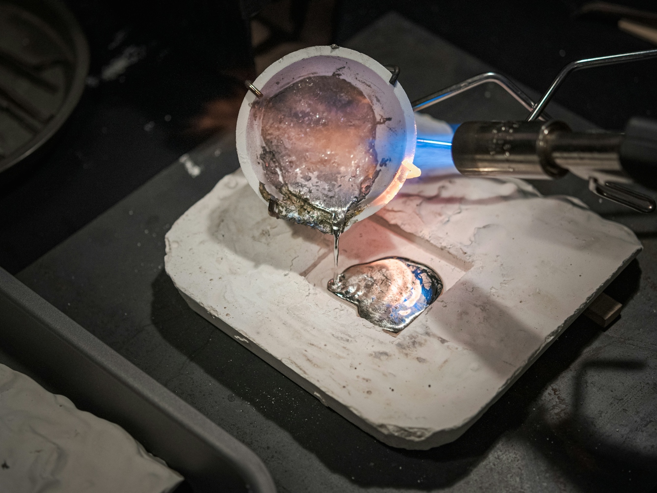 Photograph of molten lead being poured into from the ceramic heating container into the plaster mould.  A flame burner appears to the right, applying a continuous source of heat to the lead. 