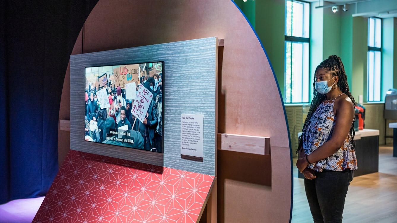 Photograph of a young woman wearing a face covering exploring an exhibition gallery space. The woman is standing to the right of frame, arms clasped across her body, reading an information panel on the front of a large circular wooden board. This text is hidden from view as the camera is looking at the back of this wooden board. Behind the board is a large TV screen mounted to a vertical rectangular board wich is covered in a blue patterned upper section and a re patterned lower section. On the screen is a still from a video showing a demonstration or protest with people carrying banner. One of the banners has the words, 'No justice, no peace, no racist police!' written on it. Behind the young woman the gallery space recedes into the distance, with further exhibits and two large windows.