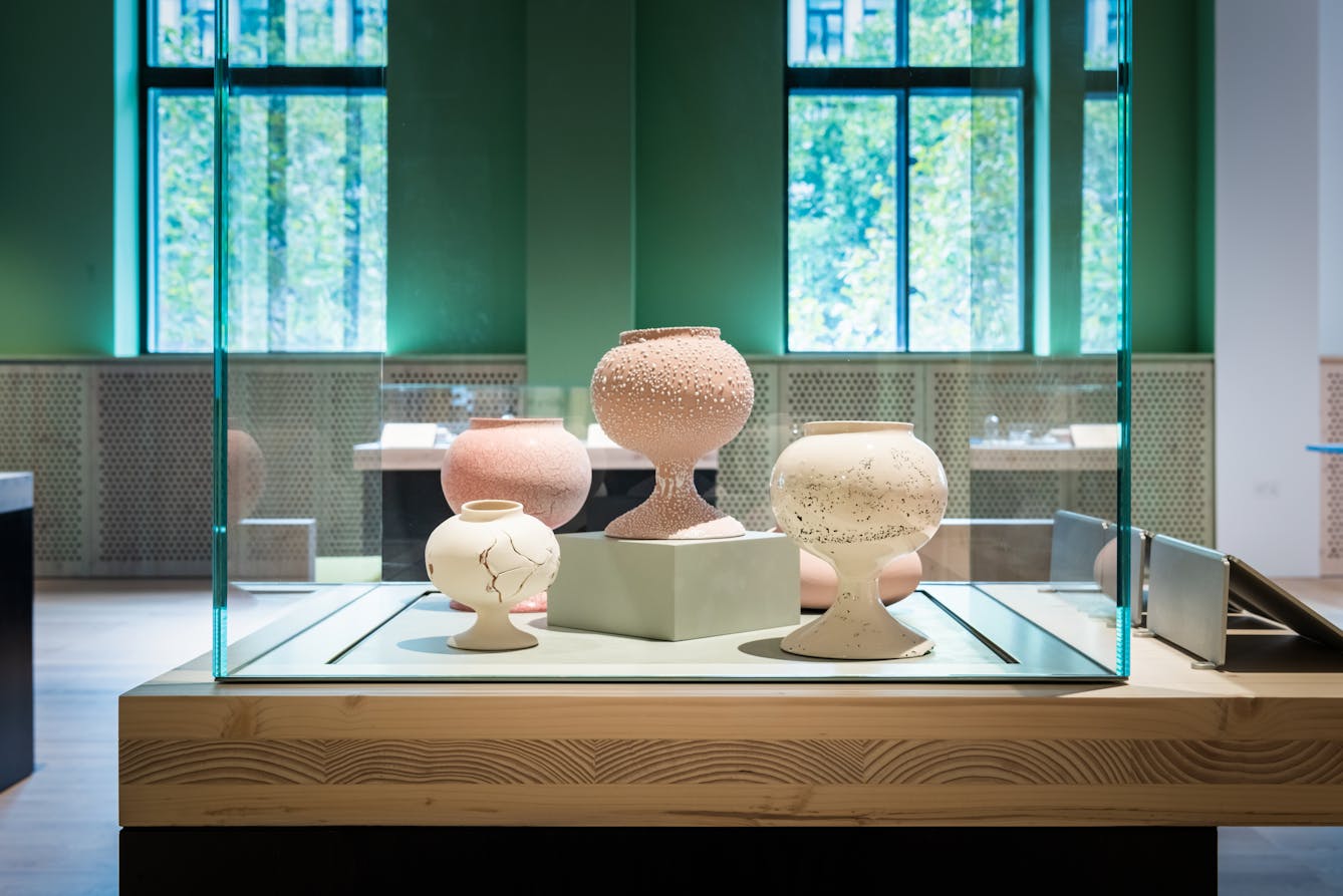 Photograph of an exhibition display case containing ceramic artworks which resemble pots or vases, coloured in light pinks and yellows. The surface of the objects are sometimes cracked, sometimes textured.