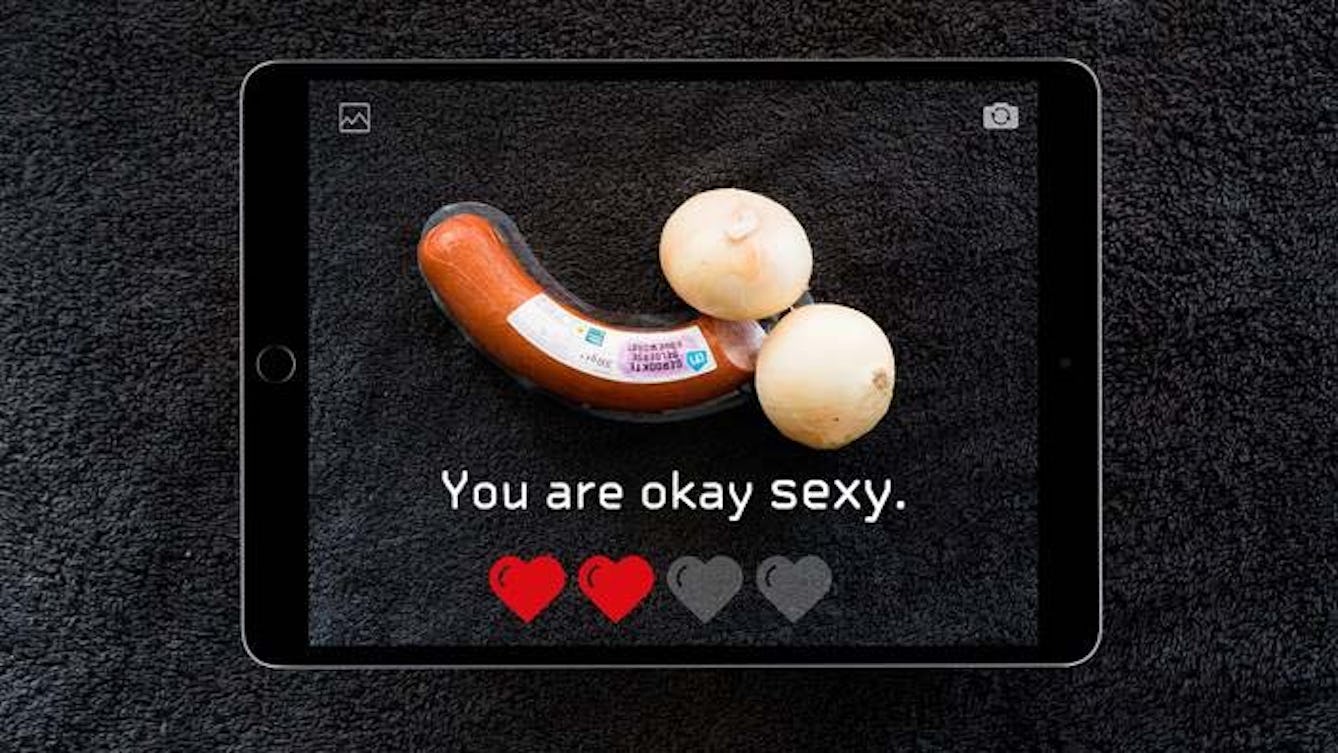 Image of an iPad taking a photo of two onions and a sausage positioned to look like a penis and testicles.  The text on the screen says "You are okay sexy." Underneath, is a rating of two red hearts out of four.