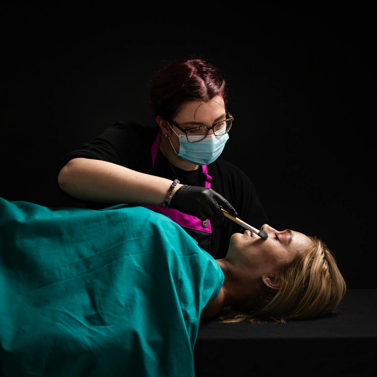 Photograph of a woman lying on her back with her eyes closed. She is covered by a green surgical sheet. Behind her stand a female makeup artist wearing a face mask and glasses. She is holding a makeup brush in her right hand, gently brushing the woman's face. her hand is wearing a black latex glove. The scene is predominantly dark black, with a hard light shining down from the top right, the source of which is out of the frame.