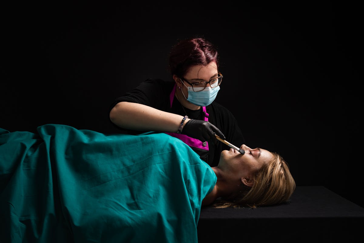Photograph of a woman lying on her back with her eyes closed. She is covered by a green surgical sheet. Behind her stand a female makeup artist wearing a face mask and glasses. She is holding a makeup brush in her right hand, gently brushing the woman's face. her hand is wearing a black latex glove. The scene is predominantly dark black, with a hard light shining down from the top right, the source of which is out of the frame.