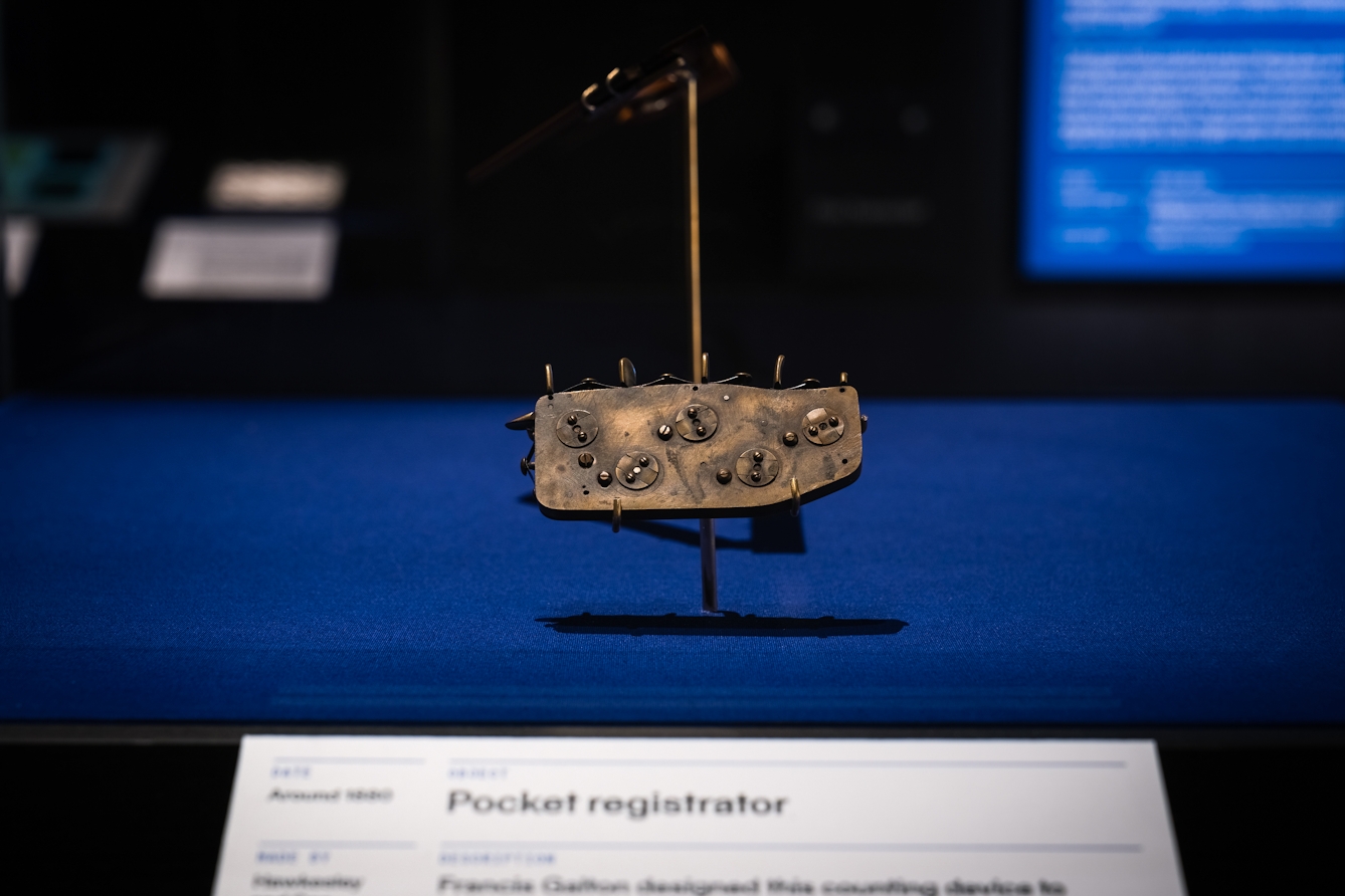 Photograph of an exhibition display case in a dark gallery space. The case contains a hand sized brass device with levers and dials, displayed suspended above a blue fabric base. In front of the case is an information panel.