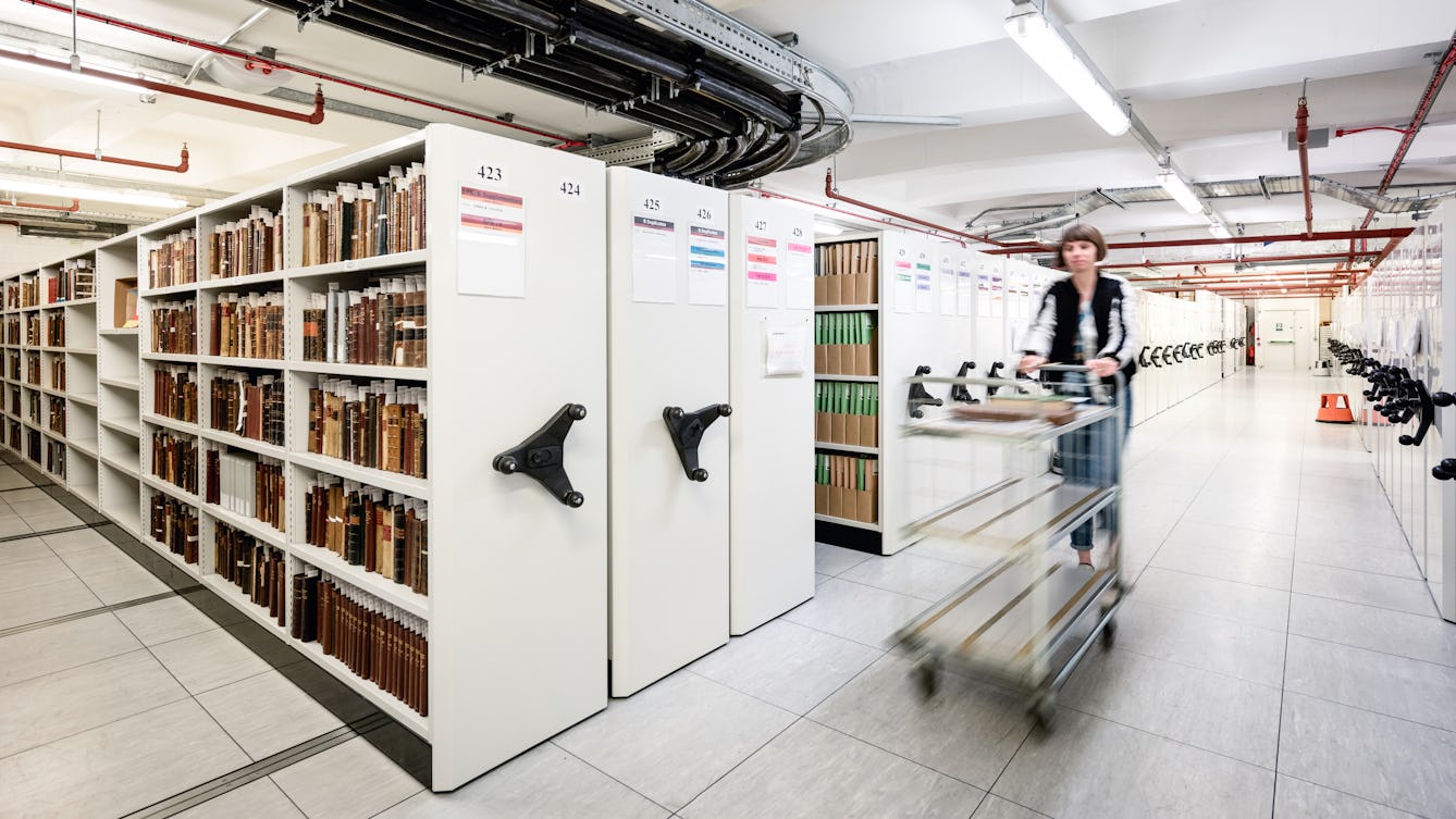 Photograph of a woman pushing a book trolley within a book store environment. She is blurred in the image due to her forward motion. She is walking down a corridor with 'rolling stack shelving' on either side. On the far left the spines of fragile books can be see, housed within the shelving.