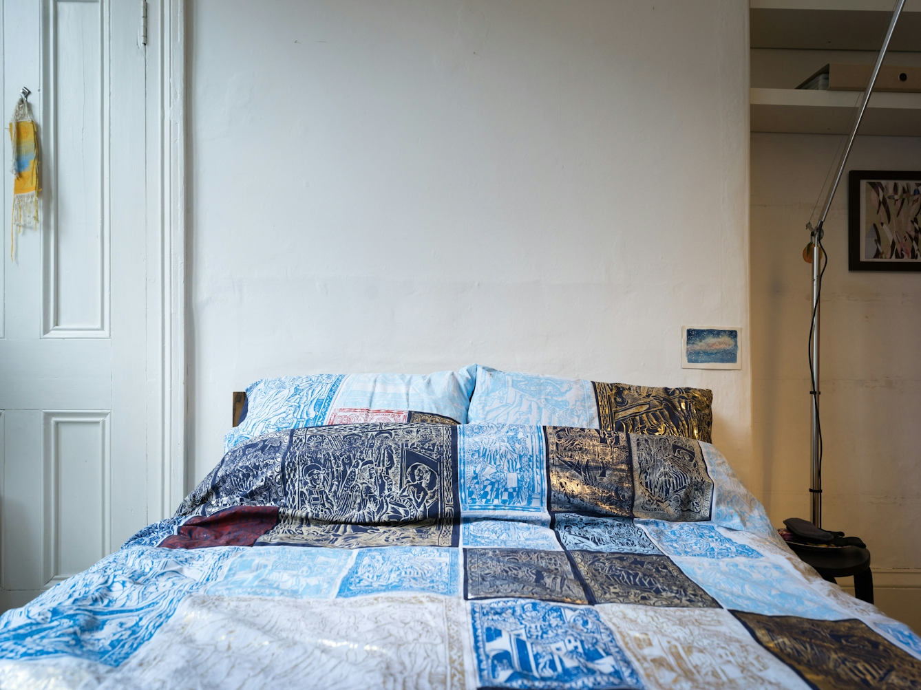 Photograph of a bedroom showing a double bed made up with a duvet cover and pillowcases which are made of a patchwork of blue, red, silver and gold screen-printed designs. To the left of the bed is part of a door. To the right is part of a lamp stand, some shelves and a framed print on the wall.