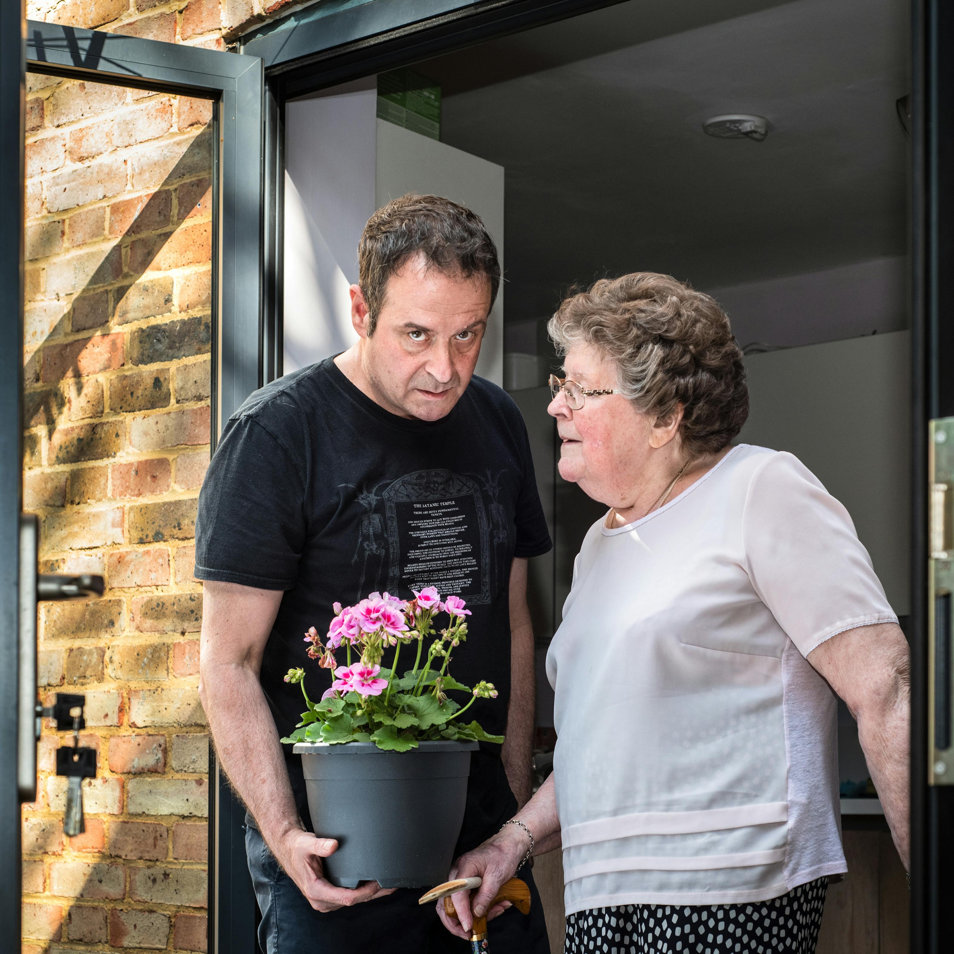 Photograph of a man and his mother stood side by side in a patio doorway. The man wearing a black t-shirt and blue trousers standing on the left is holding a plant pot containing a pink flowering plant. He is looking to camera with a long suffering look on his face. His mother, standing on the right, is wearing a pink blouse and blue patterned skirt and is looking towards him, her face in profile. Her right hand is holding the handle of her walking stick. To the left of them can be seen the patio doors with a bunch of keys in the lock and the brick wall of the back of the house. To the right of them is a section of the door frame.