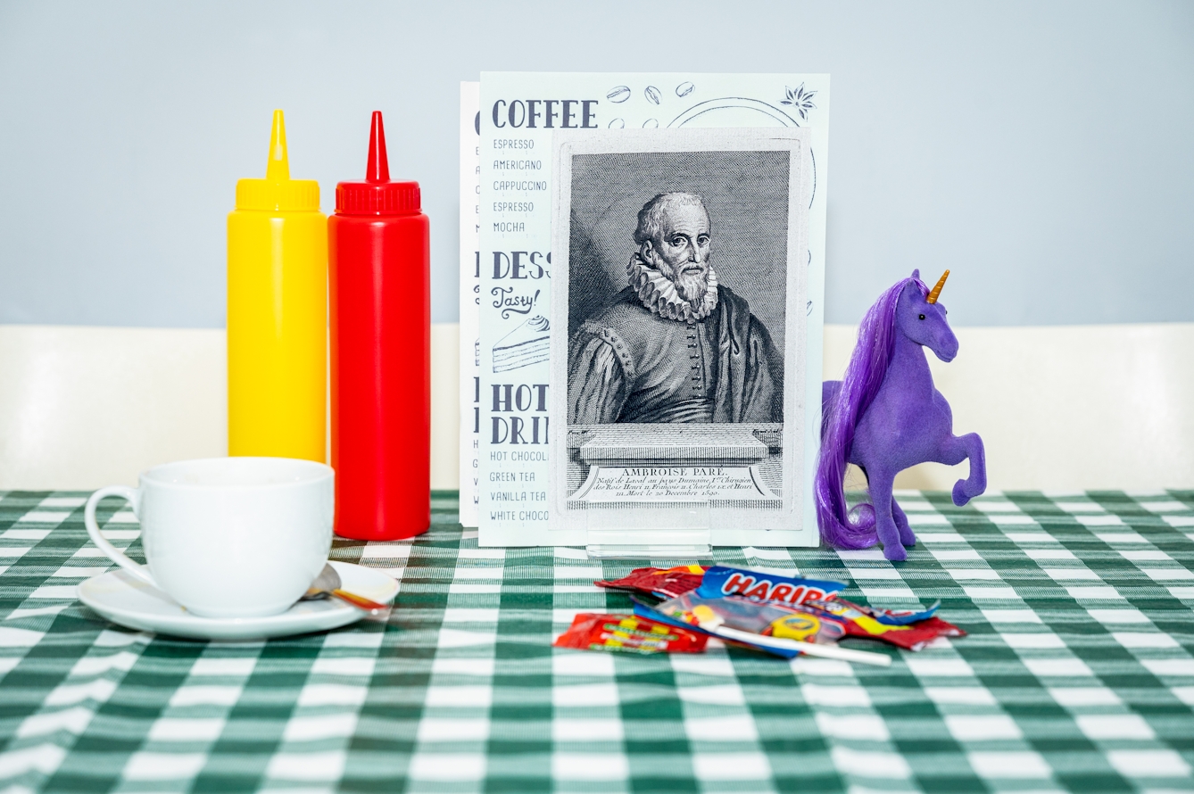 Photograph of a green checkered tablecloth in a café. On the table is a white tea cup and saucer, a collection of sweet wrappers, ketchup and mustard bottles and a menu holder with a couple of menu sheets. In front of the menus, also tucked in the menu holder is a black and white print of a man with a beard wearing a ruff, from the 16th century. Under the portrait can be seen the name Ambroise Paré. To the right of this portrait, appearing from behind it, is a purple figurine of a unicorn with an orange horn and a long purple mane.