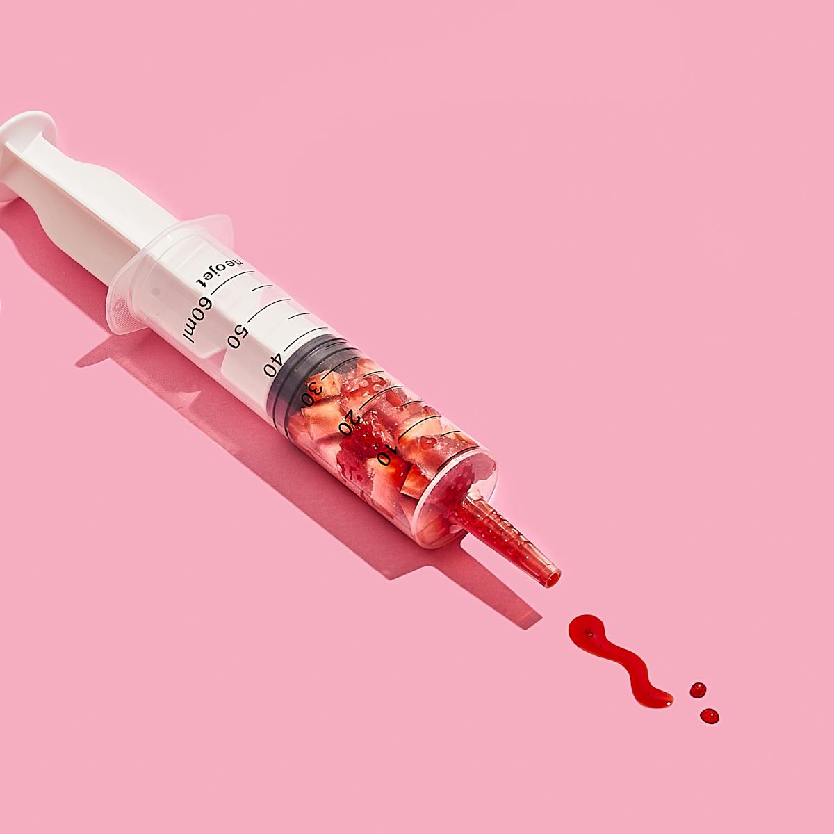 Photograph of a medical syringe lying on its side on a bright pink background. The syringe is full of chunks of cut up strawberry, compressed down into the lower half of the graduated tube. The nozzle of the syringe contains bright red strawberry juice. Some of this juice has been pushed out of the syringe and formed two dots and a squiggle on the background, reminiscent of marks made with a pen on paper. At the plunger end of the syringe are 2 strawberries lying on their side on the background.
