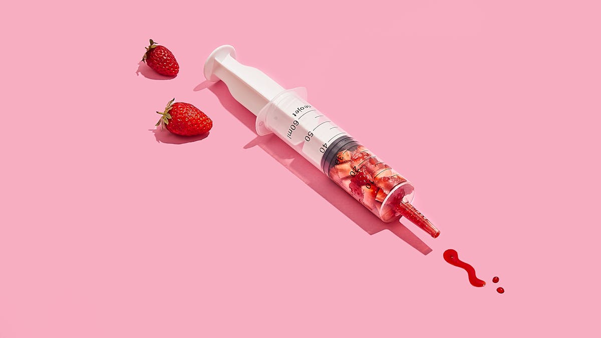 Photograph of a medical syringe lying on its side on a bright pink background. The syringe is full of chunks of cut up strawberry, compressed down into the lower half of the graduated tube. The nozzle of the syringe contains bright red strawberry juice. Some of this juice has been pushed out of the syringe and formed two dots and a squiggle on the background, reminiscent of marks made with a pen on paper. At the plunger end of the syringe are 2 strawberries lying on their side on the background.