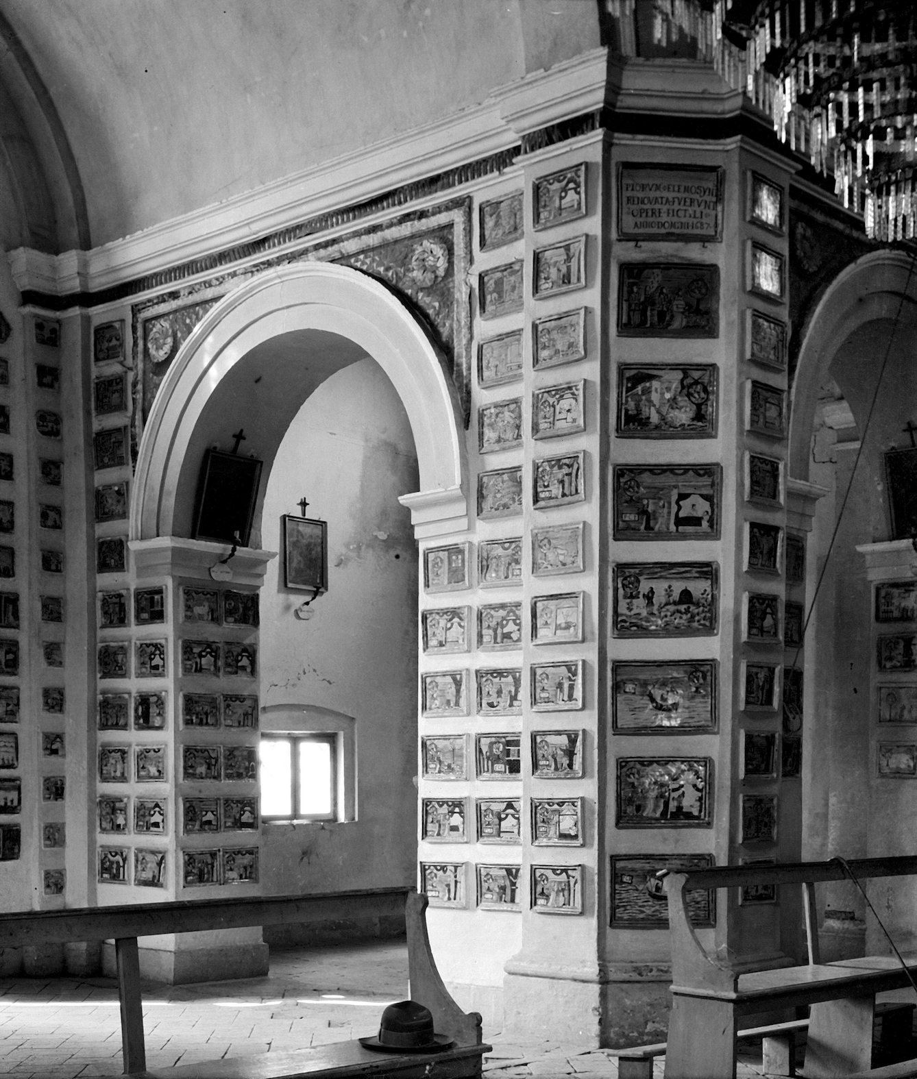 Photograph of votive tablets (ex-voto paintings) in the Church of Santa Marie de Bagni. Deruta. The image shows an archway door leading to another room and surrounding walls which are covered in votive tablets with paintings and text upon them. In front of the archway are several church pews and on the ceiling is a chandelier. 