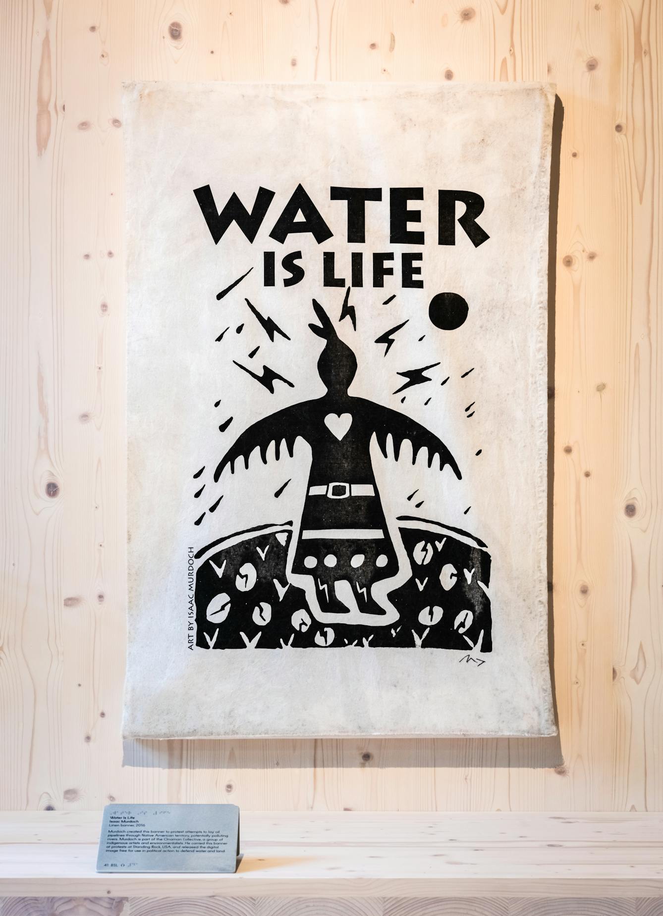 Photograph of a artwork banner hung in a gallery setting on a stained wood wall. On the banner is written the words 'water is life'. Below the words is an illustrated human figure in black and white, set in the context of land, sky and weather.
