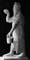 Black and white photograph of a statue depicting a man wearing sandals and a cloak that has a fish-scale texture. He is holding some sort of bucket.