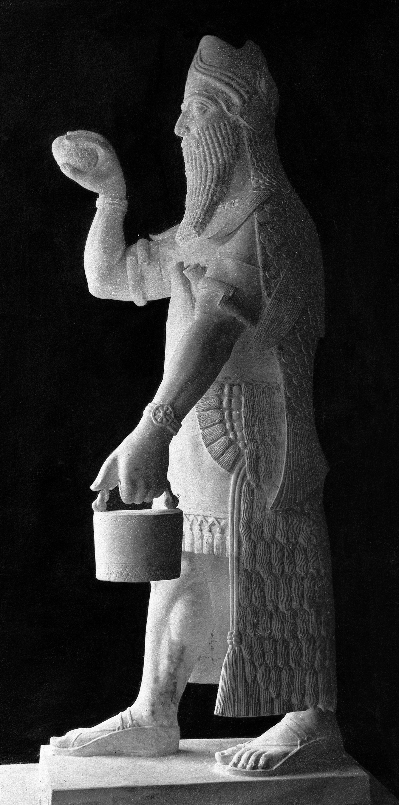 Black and white photograph of a statue depicting a man wearing sandals and a cloak that has a fish-scale texture. He is holding some sort of bucket.