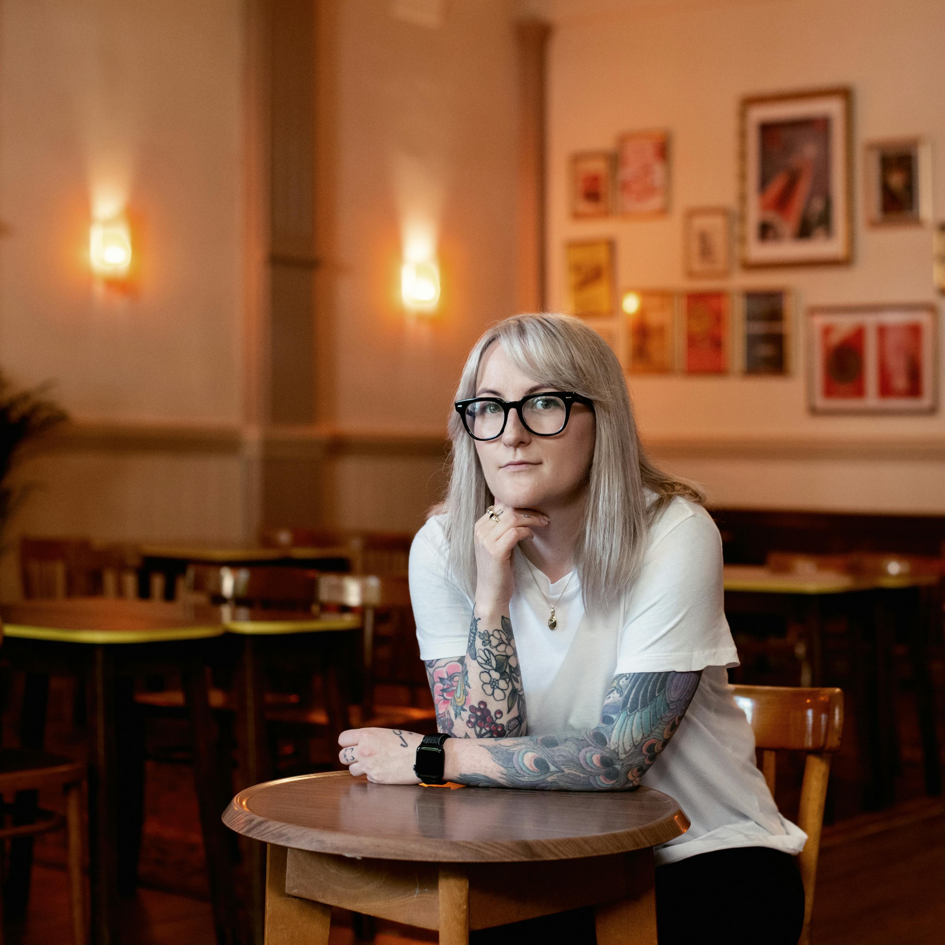 Photograph of a young woman with shoulder length blond hair seated indoors in a pub environment at a small circular wooden table. She is leaning forwards, one arm flat on the table, the other resting on its elbow, her hand tucked under her chin. She is wearing glasses looking at the camera, in a white t-shirt. Both her arms are adorned with tattoos. Behind her in the background are other empty tables in the pub and walls covered in framed pictures and a couple of wall lights.