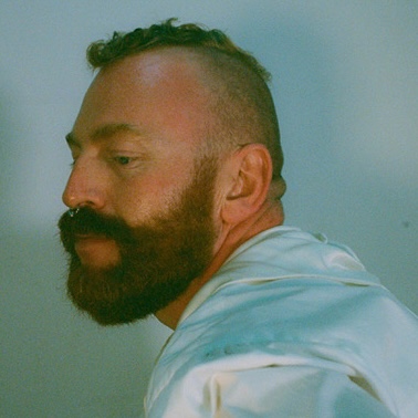 Side-profile headshot of a person with short hair and a full beard, wearing a pale shirt against a plain blue-grey background.